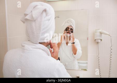 Junger Mann im weißen Morgenmantel, der sich mit Gesichtsmaske entspannt Stockfoto