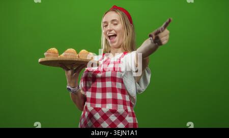 Die Chefin in roter karierter Schürze hält ein hölzernes Tablett mit Muffins und zeigt den Finger auf die Kamera im Studio; lustiges Backen. Stockfoto