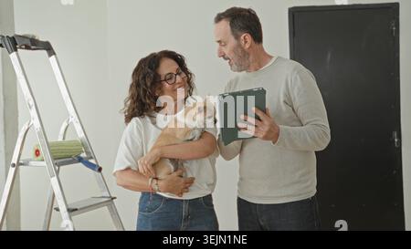 Der Mann hält ein Tablet, während die Frau einen kleinen Hund in die Wiege legt und mit der rechten Hand in Richtung Leiter und Malerwalze in einem Gebäude zeigt; Zufriedenheit bei der Renovierung des Hauses. Stockfoto