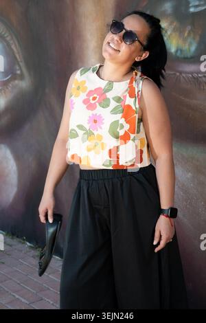 Junge Erwachsene Frau lächelt, trägt Sonnenbrille und Blumenoberteil, hält Schuhe an einem Street Art Wandgemälde Stockfoto