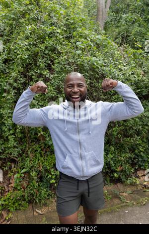 Afroamerikanischer Mann, der beide Arme auf befestigtem Weg durch dichte Sträucher in Hoodie und Shorts biegt. Fitness, Outdoor, Natur, GREE Stockfoto
