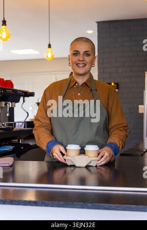 Weibliche Barista mit grüner Schürze, die Getränketräger mit zwei Deckeln an der Theke hält, lächelnd. Café, Kaffee, Espresso, Takea Stockfoto