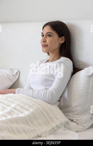 Frau, die auf dem Bett sitzt, weiß gerippte Oberseite trägt und sich an das gepolsterte Kopfteil mit weißer Decke lehnt. Weiblich, Lifestyle, minimal, Stockfoto