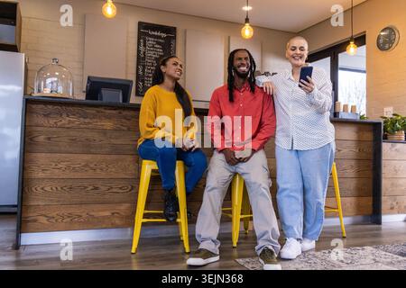 Verschiedene Freunde sitzen und lehnen sich, plaudern mit gelben Hockern und Smartphone am Café. Kaffeehaus, Bistro, Tafel Stockfoto