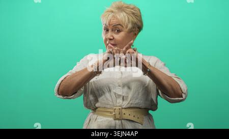 Eine ältere hispanische Frau in weißer Bluse und beigem Gürtel zeigt die Zeigefinger im Studio mit grünem Hintergrund nach oben, Hände nach vorne; eine starke Warnung. Stockfoto