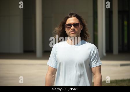 Porträt eines athletischen kaukasiers mit langen lockigen Haaren mit Sonnenbrille und hellblauem T-Shirt vor dem Bürogebäude. Stockfoto