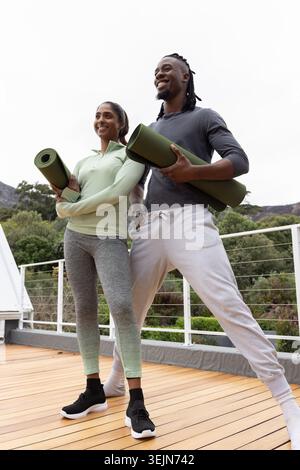 Verschiedene Freunde stehen auf einer erhöhten Holzterrasse mit gerollten grünen Yogamatten und tragen Aktivkleidung. Outdoor, Terrasse, Fitness, du Stockfoto