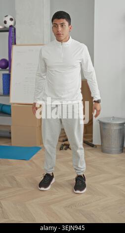 Mann, der in einem Trainingsstudio mit Yogamatte und Whiteboard mit schwarzen Sneakers und grauen Jogginghosen hockt; Konzentration Stockfoto