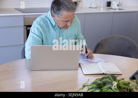 Silberner Laptop, der offen auf einem runden hölzernen Küchentisch mit gedruckten Karten und grüner Pflanze sitzt. HomeOffice, Senior, Finanzen, Plan Stockfoto