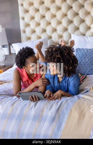 Zwei afroamerikanische Frauen, die auf dem Bett liegen, tragen ein Korallenoberteil und ein Jeanshemd und halten eine Tablette. Schlafzimmer, getuftet, Kopfteil, Kissen, Stockfoto