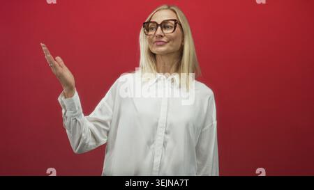 Frau zeigt mit dem Finger auf die offene Palme im roten Studio, während sie lächelt und leeren Raum präsentiert; fröhliche Demonstration. Stockfoto