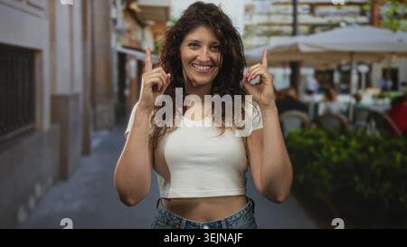 Die Frau zeigt beide Zeigefinger auf eine Straßenterrasse, lächelt mit sichtbarem Mittelriff; Freude. Stockfoto