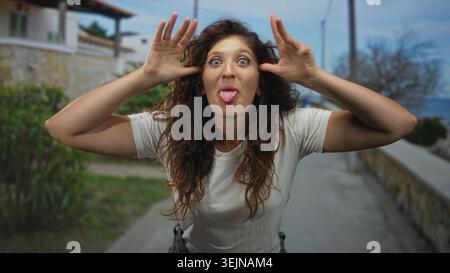 Frau mit überkreuzten Armen und sichtbaren Händen auf einer Strandpromenade in der Nähe einer Steinmauer und Sträucher; spielerisches Vertrauen. Stockfoto