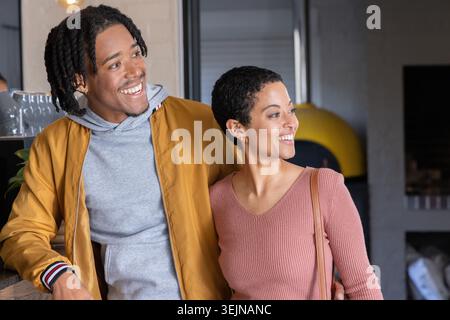 Afroamerikanisches Paar, das am Café steht, in Senfjacke, Rose Top, Tasche, lächelnd. Küche, Backofen, Gläser, Bric Stockfoto