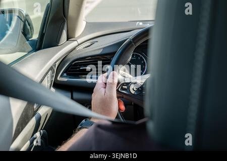 Anonymer Fahrer vom Rücksitz aus gesehen, der das Lenkrad im modernen Auto hält, der Sicherheitsgurt überkreuzt den Vorderboden, die Ansicht des Armaturenbretts und der Seitenfenster Stockfoto