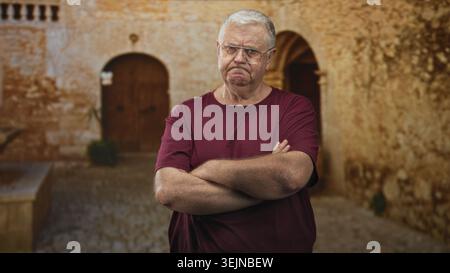 Ein älterer Mann mit überkreuzten Armen und einer Brille, die vor dem Innenhof eines alten Steingebäudes die Stirn runzelt, trägt ein kastanienbraunes Hemd und sichtbare Falten an den Unterarmen; Stockfoto