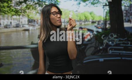 Thailändische Frau hält ein weißes Puzzleteil mit der Hand nahe ihrem Gesicht auf einer Straße an einem Kanal mit Fahrrädern und Bäumen; Neugier. Stockfoto