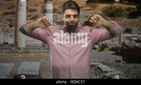 Hispanischer Mann mit Bart zeigt Daumen mit beiden Händen vor dem alten Gebäude aus Steinruinen; Kritik missbilligt. Stockfoto