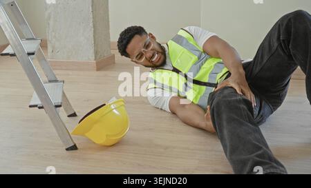 Mann, der ein verletztes Bein neben gefallenem Schutzhelm und Leiter auf einem Bauboden in einem Gebäude festhält; Unfallschmerz. Stockfoto