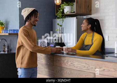 Afroamerikanischer Gast und Barista tauschen To-go-Tassen am Kaffeeschalter mit POS-Schild auf der Schürze aus. Kaffee, zum Mitnehmen, gemütlich, mod Stockfoto