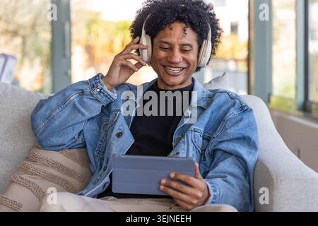 Afroamerikaner sitzt zu Hause auf dem Sofa, trägt Denimjacke und Kopfhörer und hält ein Tablet. Sofa, Fenster, Tageslicht, neu Stockfoto