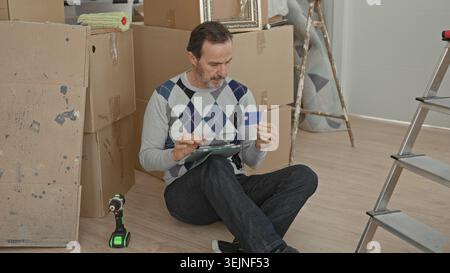 Mann, der eine Kreditkarte hält und ein Tablet anzapft, während er zwischen Kartonkartons und Leiter in einem Gebäude sitzt; fokussierte Planung. Stockfoto