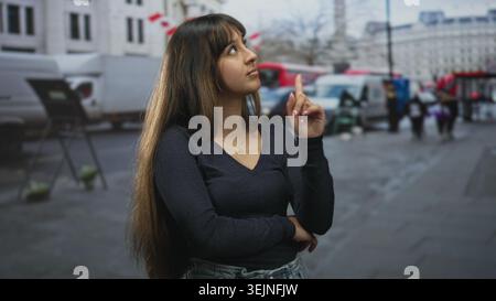 Die junge hispanische Frau zeigt den Zeigefinger nach oben mit gefaltetem Arm und sichtbarer Halskette auf der Straße in der Nähe von geparkten Lieferwagen und Fußgängern; ruhige Besinnung. Stockfoto