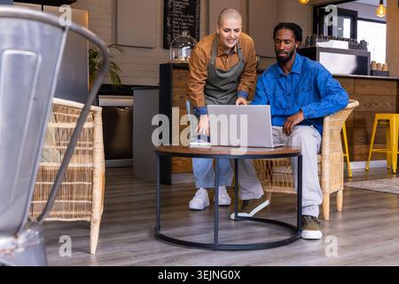 Weibliche Barista, die eine Schürze trägt und auf den Laptop zeigt, während ein afroamerikanischer Mann im Café zusieht. Café, Zusammenarbeit, Teammitglied Stockfoto