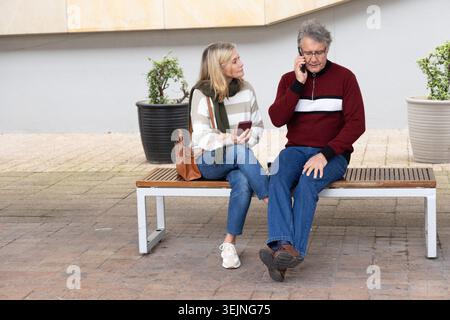 Ein Paar mit einem älteren Mann, der auf der Bank auf dem plaza sitzt, Smartphones benutzt, braune Ledertasche, Pflanzgefäße. Urban, Outdoor, Duo, Freizeit, P Stockfoto