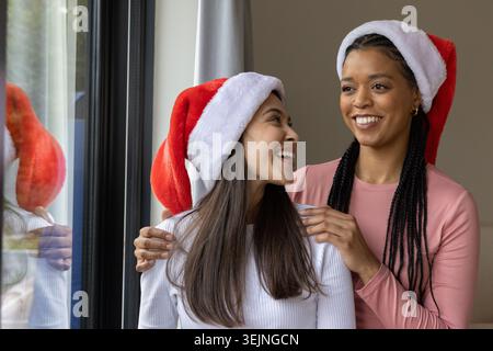 Verschiedene weibliche Freunde stehen zu Hause neben der Glastür, tragen Santa-Hüte, lachen, umarmen. Duo, lachen, festlich, Fenster Stockfoto