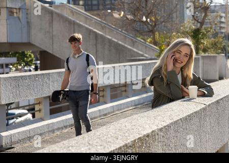 Frau lehnt sich auf Rampengeländer und spricht am Telefon mit Tasse, während Mann Skate Rucksack-Headset läuft. Stadtverkehr, Überführung, Beton, Stockfoto