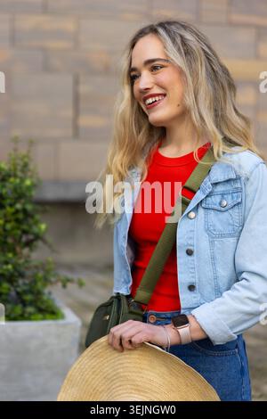 Lächelnde Frau, auf dem platz neben der Steinmauer stehend, mit Strohhut und Jeansjacke. Outdoor, Urban, Freizeit, Crossbody, Stockfoto