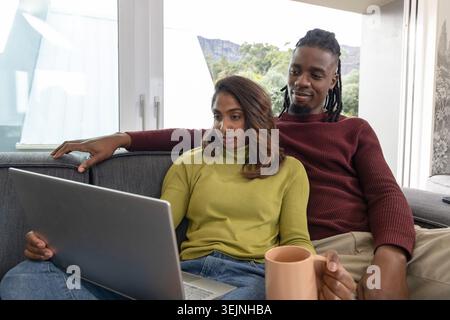 Verschiedene Paare in olivfarbenen, burgunderfarbenen Pullover sitzen zu Hause auf grauem Sofa und sehen einen Laptop mit Becher. Duo, gemütlich, innen, taglig Stockfoto