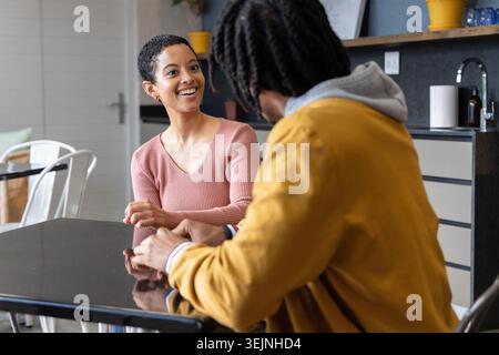 Afroamerikanische Freunde plaudern und Gesten an einem glänzenden schwarzen Tisch in der Küche, Frau in rosa Spitze. Konversation, zwanglos, sozial Stockfoto