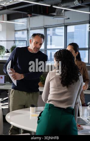 Verschiedene Kollegen, die gedruckte Karten um den runden weißen Tisch im Großraumbüro mit Laptop überprüfen. Teamwork, Diagramme, Analysen, modern, Urban, funktioniert Stockfoto