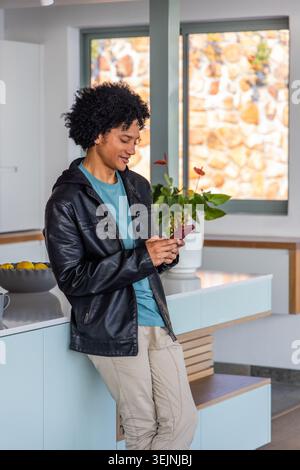 Afroamerikaner 20iger, der sich in schwarzer Jacke mit Smartphone auf die Kücheninsel lehnt, Kopierraum. Telefon, Lifestyle, modern, interi Stockfoto