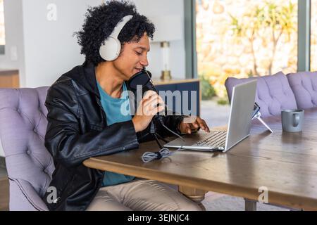 Afroamerikanischer Mann mit Kopfhörern und Jacke, der am Esstisch mit Laptop ins Mikrofon spricht. Podcast, Mikrofon, Computer Stockfoto