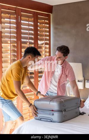 Zwei männliche Freunde stehen am Bett und packen graue Hartschalenkoffer in der Nähe von Holzläden. Begleiter, Reise, Gepäck, Hotel, Betro Stockfoto