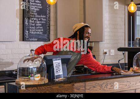 Afroamerikanischer männlicher Barista in grüner Schürze, lehnt sich an der Theke, Café-Bar, Wischtuch in der Nähe von POS. Café, Espresso, Tafel, Stockfoto