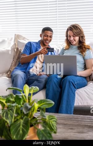 Ein Paar sitzt auf dem Sofa und ein afroamerikanischer Mann zeigt auf die Kreditkarte, während eine Frau einen Laptop hält. Duo, Computer, Zuhause, gemütlich, Kissen, Jalousien, Pflanze Stockfoto