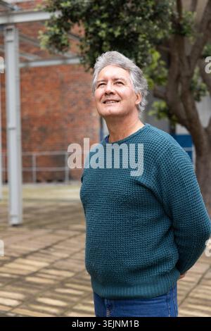 Ein älterer Mann steht im gepflasterten Innenhof, trägt einen blauen Strickpullover und eine blaue Jeans und blickt nach oben. Älter, Gentleman, draußen, urb Stockfoto