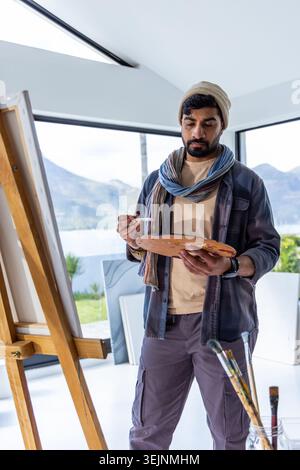 Indischer Mann in den dreißiger Jahren, der im Studio an der Staffelei stand, Hut trug, Palette hielt und den Blick auf den See sah. Künstler, Pinsel, Leinwand, PA Stockfoto