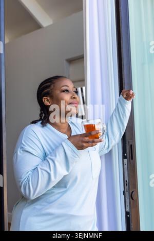Afroamerikanische Frau, die in hellblauer Loungewear an der Schiebetür steht und eine Tasse Tee hält. Zöpfe, entspannt, Morgen, Su Stockfoto