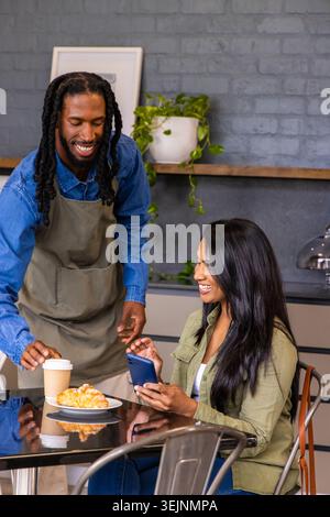 Afroamerikanischer Barista in Denim-Schürze überreicht Kaffeetasse an indische Gäste mit Telefon im Café. Becher, Croissant, Lächeln, Interac Stockfoto