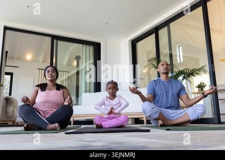 Afroamerikanische Familie sitzt im Kreuz auf Yogamatten im Wohnzimmer und meditiert in Activewear. Trio, Achtsamkeit, Gelassenheit, Stockfoto