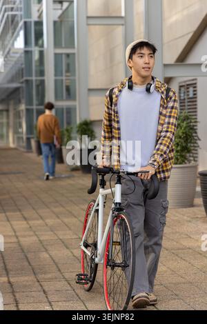 Asiatischer Mann schiebt weißes Rennrad mit roten Felgen, während er Mütze und Kopfhörer auf dem Flur trägt. Urban, Fahrrad, Pendler, Cycli Stockfoto