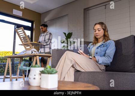 Ein Paar sitzt zu Hause, eine Frau liest Tablette in einer Jeansjacke auf dem Sofa, während ein Mann einen Stuhl zusammenstellt. Wohnzimmer, sonnenbeleuchtet, modern, cas Stockfoto