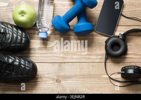 Sport- und Fitnessgeräte mit Turnschuhen, Kurzhanteln, Handtuch und Kopfhörern auf Holzbrett. Draufsicht, Platz für Ihren Text. Stockfoto