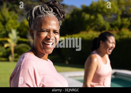 Verschiedene Freundinnen stehen in der Nähe des Swimmingpools auf dem Rasen und tragen hellrosa Oberteile. Im Freien, im Garten, sonnig, lachen, Stockfoto