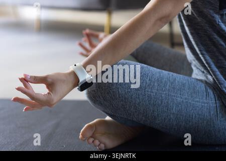 Erwachsene Frau sitzt im Kreuz auf dunkler Yogamatte zu Hause, trägt graue Leggings und weiße Smartwatch. Meditation, Achtsamkeit, Ser Stockfoto